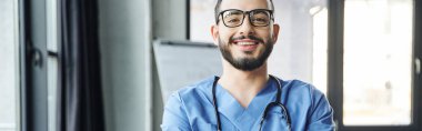 Modern klinikteki kameraya bakan sakallı, gözlüklü ve steteskoplu genç ve neşeli sağlık görevlisinin portresi, ilk yardım semineri ve acil durum hazırlık konsepti, afiş