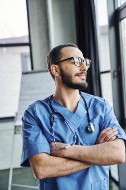 Gözlüklü, mavi üniformalı, gözlüklü, gülümseyen profesyonel sağlık görevlisi. Kollarını kavuşturup eğitim odasına bakıyor. İlk yardım semineri ve acil durum hazırlık konsepti.
