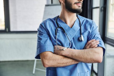 Mavi üniformalı profesyonel sakallı tıp eğitmeninin kolları çapraz ve steteskopla hastanede durması, ilk yardım semineri ve acil durum hazırlık konsepti.