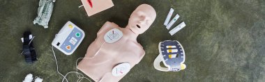 top view of automated external defibrillator, neck brace, syringes, compression tourniquet and bandages near CPR manikin, medical equipment for first aid training and skills development, banner clipart