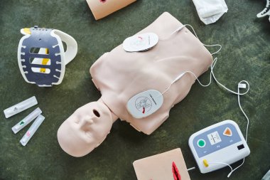 top view of CPR manikin near automated defibrillator, wound care simulators, neck brace and syringes on floor in training room, medical equipment for first aid training and skills development clipart