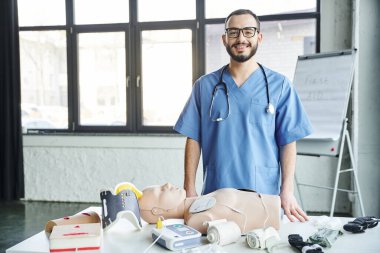 CPR manikin, defibrilatör, sıkıştırıcı bandaj ve turnike, ilk yardım öğrenme ve kritik beceri geliştirme konsepti yakınlarındaki kameraya bakan neşeli tıp eğitmeni.