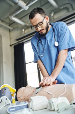 Gözlüklü ve mavi üniformalı sakallı profesyonel sağlık görevlisi kalp masajı için defibrilatör ve sıkıştırıcı bandaj yanında göğüs masajı yapıyor. Kritik beceri geliştirme konsepti.