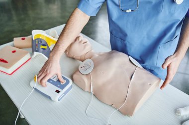 partial view of medical instructor operating automated defibrillator near CPR manikin, wound care simulators and neck brace, first aid hands-on learning and critical skills development concept clipart