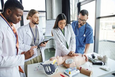 young asian woman in white coat doing chest compressions on CPR manikin near instructor, medical equipment and multicultural students with notebooks, emergency situations response concept clipart