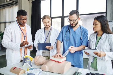 multicultural students in white coats writing on clipboards and looking at medical instructor tamponing wound on simulator near CPR manikin and medical equipment, life-saving skills concept clipart