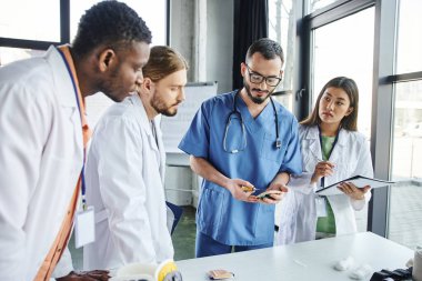 professional paramedic practicing with injection pad near diverse group of multicultural students in white coats during medical seminar in training room, skills development concept clipart