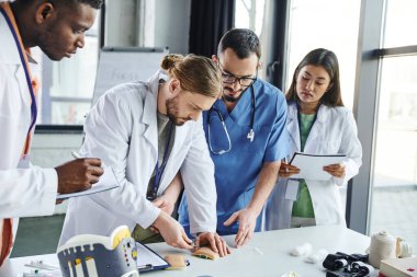İlk yardım semineri sırasında eğitim sahasında tıbbi ekipmanların yanında enjeksiyon yapan genç öğrenci, sağlık çalışanı ve çok ırklı beyaz önlüklü takım, beceri geliştirme konsepti