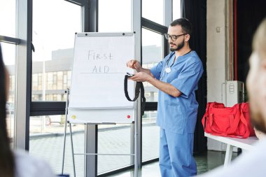 paramedic in eyeglasses and blue uniform standing at flip chart with first aid lettering and showing compressive tourniquet to students on blurred foreground, life-saving skills concept clipart