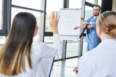 İlk yardım semineri, acil durum hazırlığı ve hayat kurtarma becerileri konsepti boyunca sıkıştırıcı turnike uygulayan ve öğrencilerin soru sormasına bakan bir tıp öğretmeni.