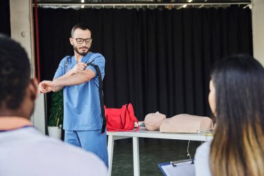 first aid seminar, medical instructor in blue uniform and eyeglasses applying compression tourniquet near interracial participants on blurred foreground, acquiring and practicing life-saving skills concept clipart
