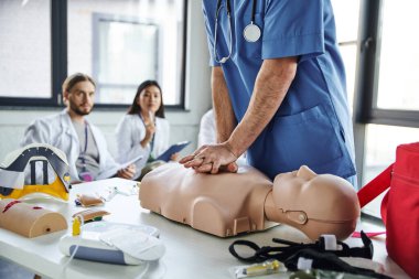 professional paramedic in blue uniform doing chest compressions on CPR manikin near medical equipment and young multiethnic students on blurred background, practicing life-saving skills concept clipart