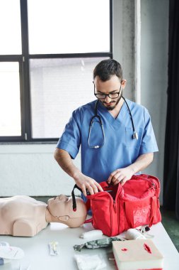 Gözlüklü ve mavi üniformalı genç tıp eğitmeni ilk yardım semineri için eğitim odasını hazırlıyor ve suni teneffüs manikinin yanındaki kırmızı çantayı açıyor. Hayat kurtaran beceri geliştirme konsepti.