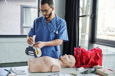 İlk yardım seminerine hazırlanırken, mavi üniformalı ve gözlüklü profesyonel sağlık görevlisi, kalp masajı manikini ve kırmızı çantanın yanında boyunluk tutuyor. Hayat kurtaran beceriler geliştirme konsepti.