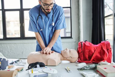 young professional paramedic practicing chest compressions on CPR manikin near red first aid bag, automated defibrillator and medical devices in training room, life-saving skills development concept clipart