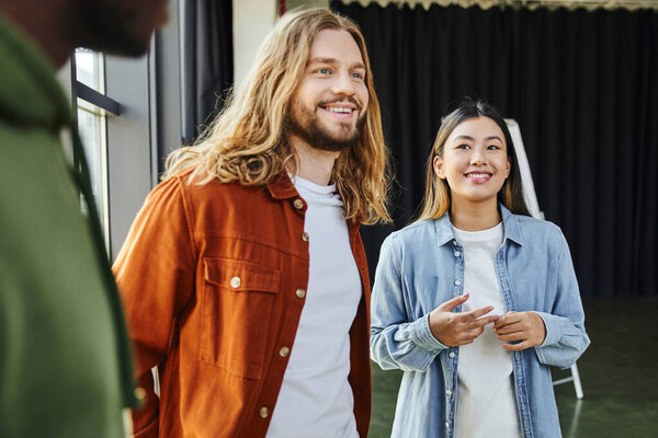 cheerful and trendy long haired man smiling near young asian woman and african american colleague on blurred foreground in modern office, successful collaboration, creative startup