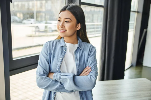 快乐的亚洲女人 一头乌黑的头发 两臂交叉地站在一起 望着窗外的现代写字楼 蓝色斜纹棉布衬衫 快乐的微笑 年轻而成功的企业家 — 图库照片