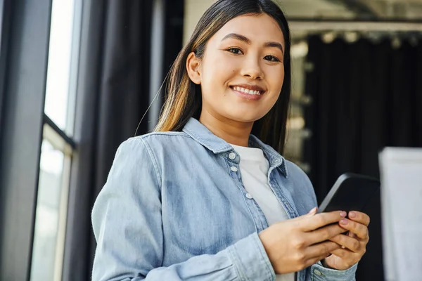 stock image youthful and trendy asian businesswoman with brunette hair and happy smile networking on mobile phone and looking at camera in contemporary office, ambitious and career oriented person