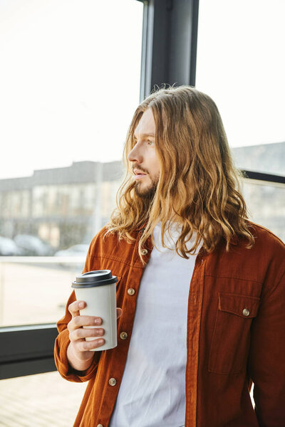 dreamy, long haired and bearded entrepreneur in casual clothes holding paper cup with coffee, standing near window and looking away, cool hipster style man in modern office