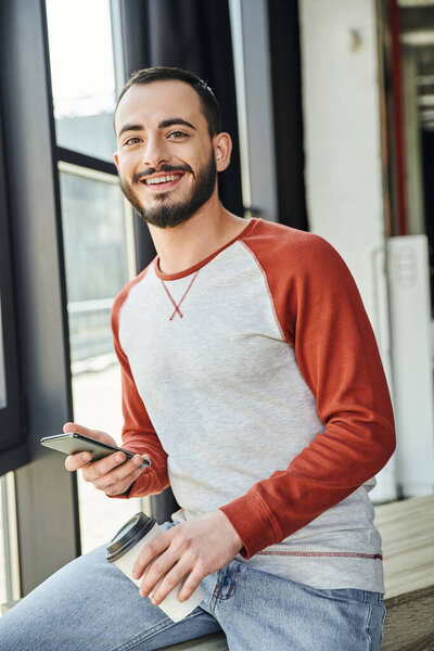 self-assured and stylish bearded man in casual clothes smiling at camera while holding mobile phone and paper cup with takeaway drink in contemporary office, young businessman 