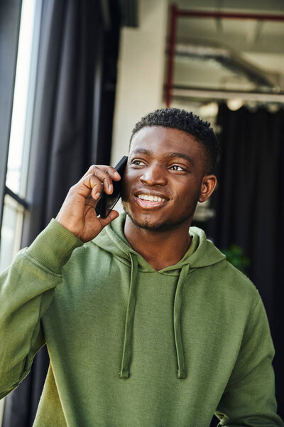 optimistic african american man in green hoodie talking on mobile phone and looking away in contemporary office, successful entrepreneur discussing business startup 
