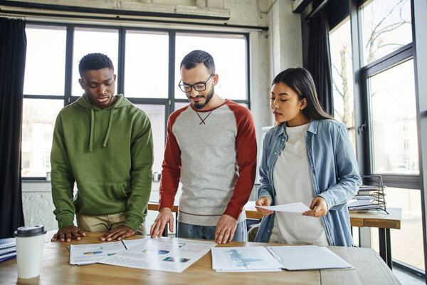 multicultural entrepreneurs in casual clothes looking at documents with graphs on desk in modern office, young and creative team working on startup planning, successful partnership