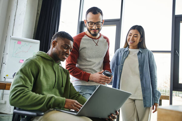smiling african american businessman working on laptop near asian woman and bearded colleague with eyeglasses and coffee to go in modern office, successful youth 