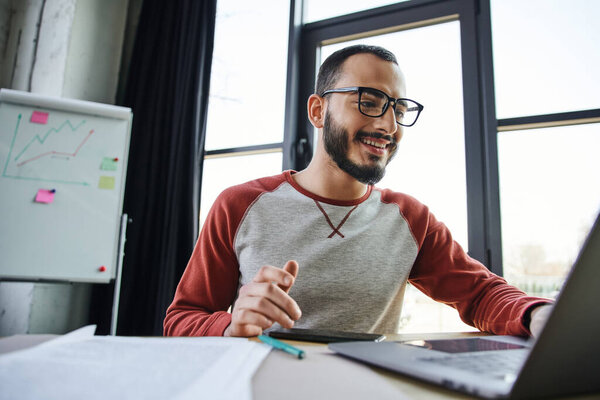 cheerful bearded businessman in eyeglasses and casual clothes working on laptop near documents and flip chart with graphs in contemporary office, young successful entrepreneur 