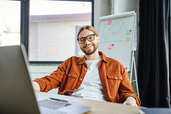 stock image happy and optimistic bearded businessman in eyeglasses and trendy shirt sitting at laptop and looking at camera next to flip chart on blurred background in contemporary office
