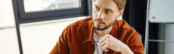 stock image portrait of thoughtful and bearded entrepreneur in trendy casual shirt holding eyeglasses and looking away while working in contemporary office, business lifestyle, banner