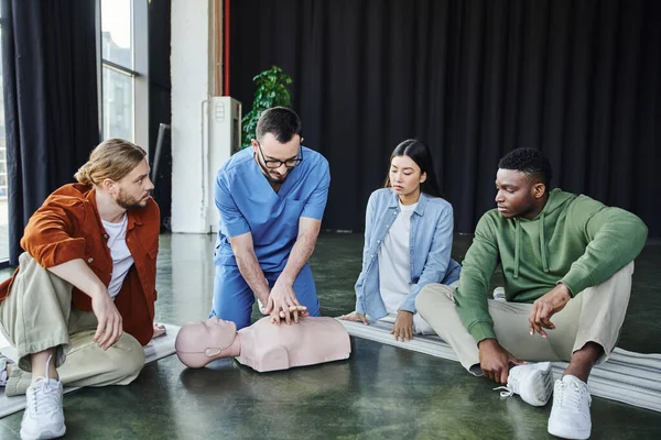 professional paramedic doing chest compressions on CPR manikin while showing cardiopulmonary resuscitation techniques to young multicultural team during first aid seminar in training room
