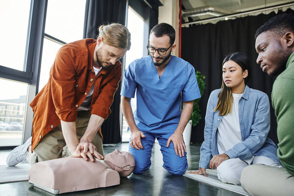 young man practicing cardiopulmonary resuscitation on CPR manikin near healthcare worker and multicultural participants during first aid training seminar, life-saving skills and techniques concept