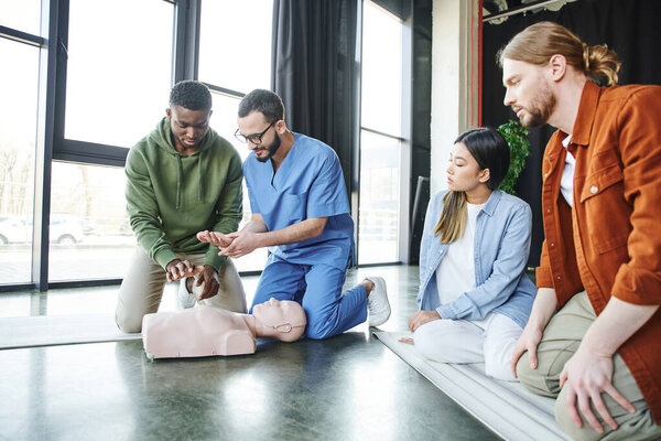medical instructor explaining african american man cardiopulmonary resuscitation techniques near CPR manikin and young multiethnic team, life-saving skills and emergency preparedness concept