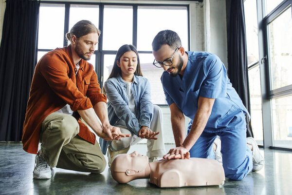 professional paramedic in eyeglasses and uniform showing chest compressions on CPR manikin near young man and asian woman during first aid training seminar, effective life-saving skills concept