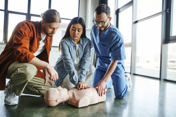 asian woman practicing chest compressions on CPR manikin near young man and medical instructor in eyeglasses and uniform during first aid seminar, emergency preparedness concept