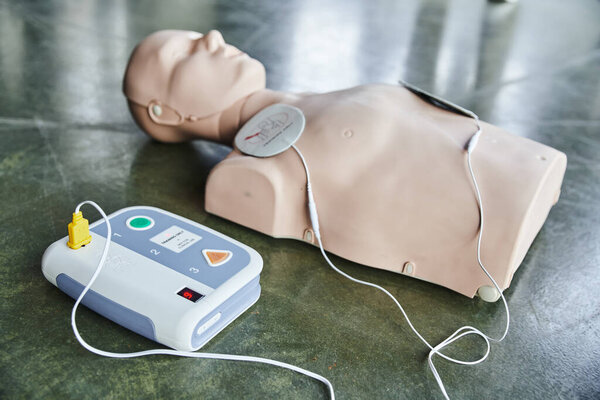 selective focus of automated external defibrillator near cardiopulmonary resuscitation training manikin on blurred background on floor in training room, medical equipment for first aid training 