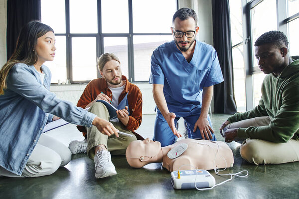 young asian woman and medical instructor pointing at CPR manikin with defibrillator near multicultural participants of first aid training seminar, health care and life-saving techniques concept