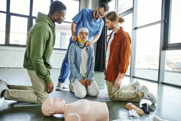 first aid training, interracial men looking at medical instructor putting neck brace on asian woman with bandaged head near CPR manikin and medical equipment, emergency situations response concept