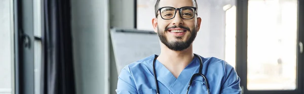 portrait of young and joyful healthcare worker with beard, eyeglasses and stethoscope looking at camera in modern clinic, first aid training seminar and emergency preparedness concept, banner