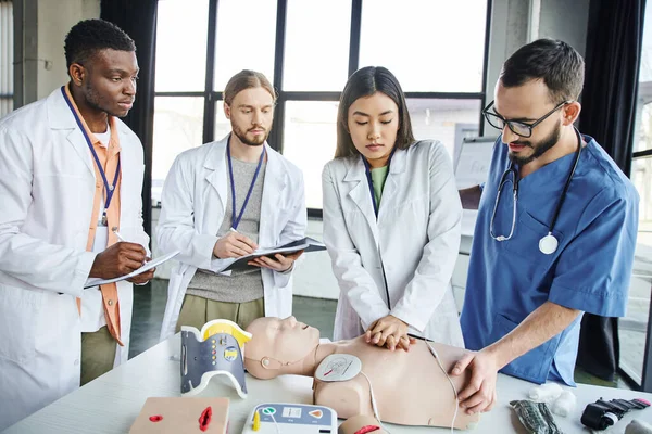 asian woman practicing chest compressions on CPR manikin near medical instructor and interracial students writing in notebooks during first aid seminar, emergency situations response concept