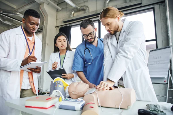 young man practicing chest compressions on CPR manikin near medical equipment, professional paramedic and interracial students writing in notebooks, emergency situations response concept