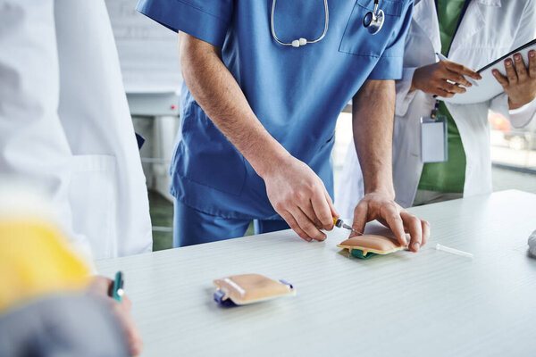 first aid hands-on learning, cropped view of medical instructor in uniform practicing with injection training pad near students in white coats, life-saving skills development concept
