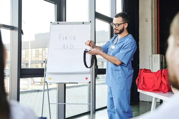 stock image paramedic in eyeglasses and blue uniform standing at flip chart with first aid lettering and showing compressive tourniquet to students on blurred foreground, life-saving skills concept