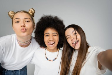 Selfie, beyaz tişörtlü, mutlu ve etnik çeşitliliğe sahip genç kız arkadaşların portresi dudaklarını bükerek ve dillerini uzatarak gri, genç arkadaşlarıyla birlikte eğlenirken ve yalnız poz verirken.