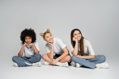 Beyaz tişörtlü ve şık kot pantolonlu neşeli ve etnik çeşitliliğe sahip genç kız arkadaşlar gri arka planda otururken ve zaman geçirirken birbirlerine bakmıyorlar.