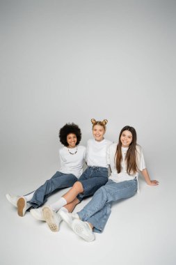 Cheerful and multiethnic teenage girlfriends in trendy jeans and white t-shirts looking at camera while sitting and posing on grey background, multiethnic teen models concept clipart