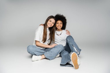 Beyaz tişörtlü, kot pantolonlu gülümseyen esmer kız birlikte oturup gri arka plandaki kameraya bakarken Afrikalı Amerikalı kız arkadaşına sarılıyor.