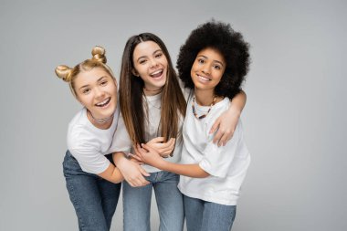 Positive multiethnic teen girls in white t-shirts and jeans hugging brunette friend and looking at camera while standing isolated on grey, lively teenage girls concept, friendship and bonding clipart
