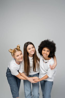 Beyaz tişörtlü ve kot pantolonlu neşeli ve etnik çeşitliliğe sahip genç kızlar esmer arkadaşa sarılıyorlar ve gri, canlı genç kızlar konsepti, arkadaşlık ve kaynaşma üzerine izole bir şekilde duruyorlar.