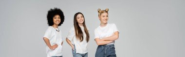 Beyaz tişörtlü neşeli ve etnik çeşitlilikteki genç kız arkadaşlar, gri, canlı genç kızlar konsepti, afiş ve pankartta yan yana dururken kameraya bakıp poz veriyorlar. 
